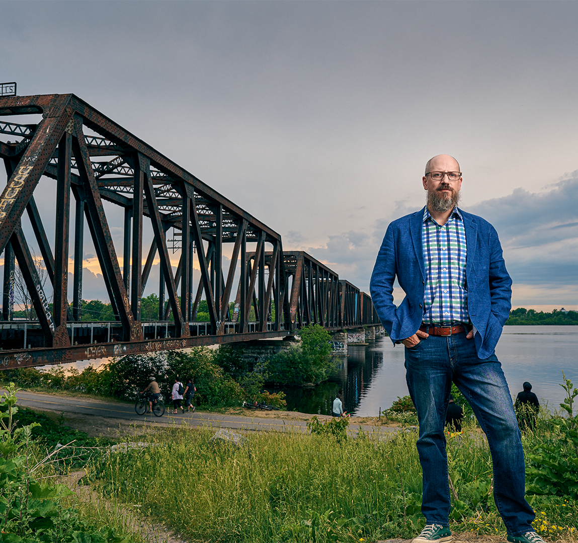 Portrait of Michael Spratt standing on grass in front of industrial bridge and water