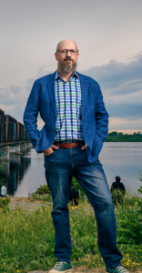 Portrait of Michael Spratt standing on grass in front of industrial bridge and water
